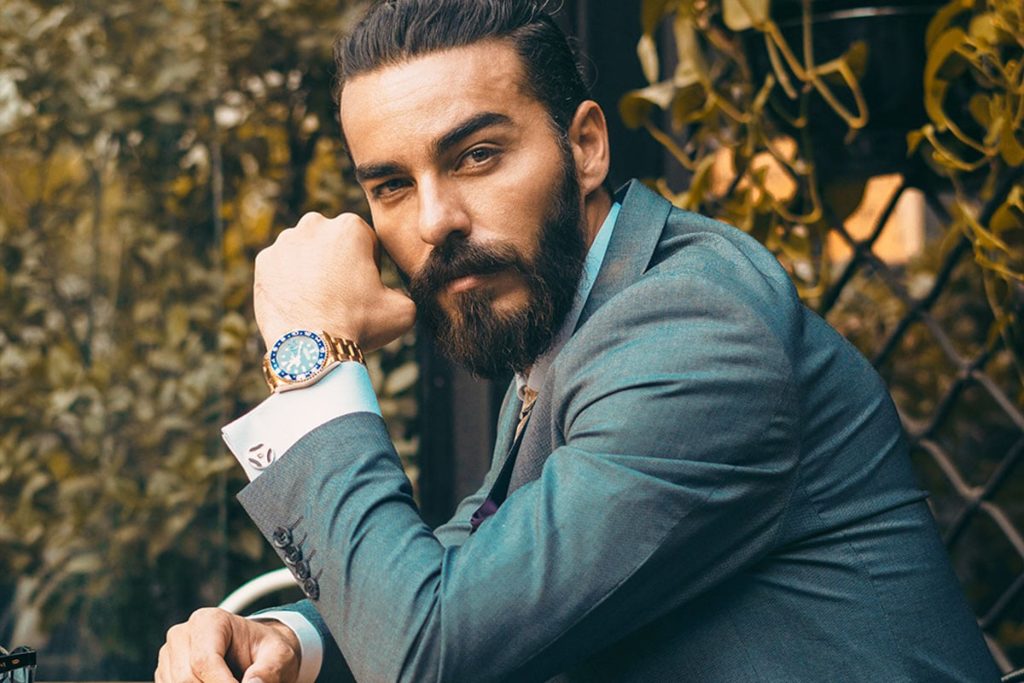 Man with well groomed beard posing with fist to cheek, showing fashionable watch