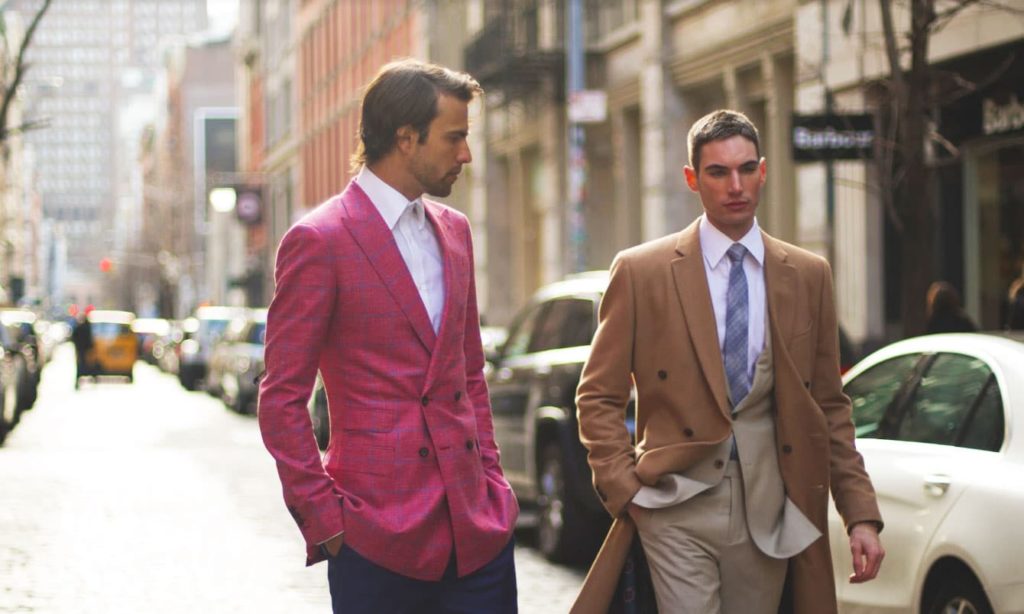 two men in different styles of custom suits. One is wearing an overcoat.