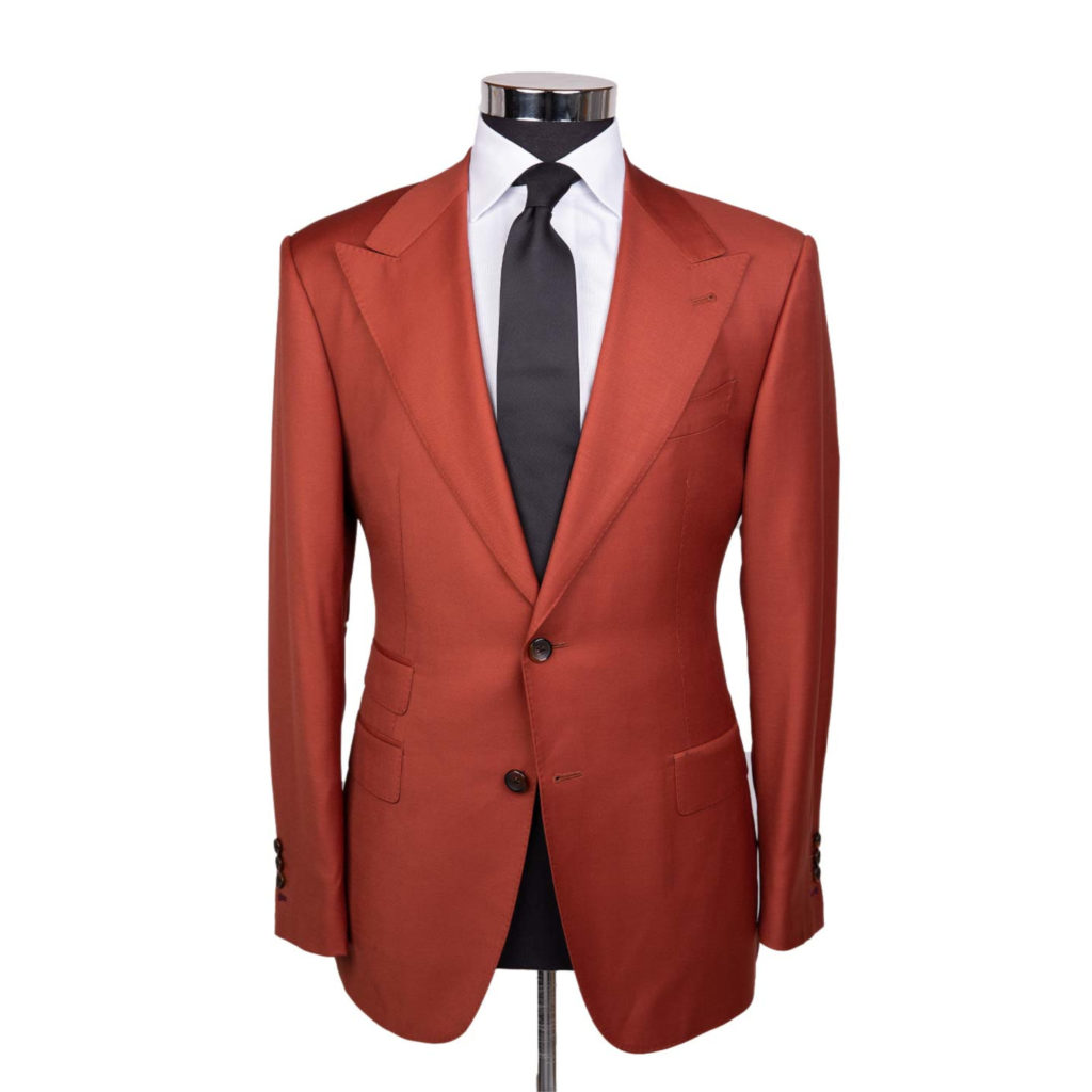 A blood orange colored suit jacket on a body form with a white shirt and a black tie on a white background