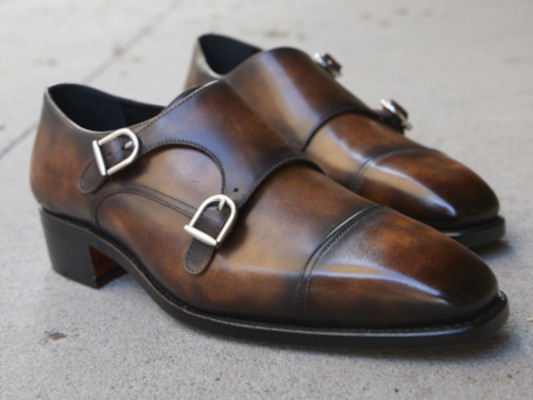 Double monk shoes in brown patina with a brass buckle. The pair of shoes is on a cement floor.