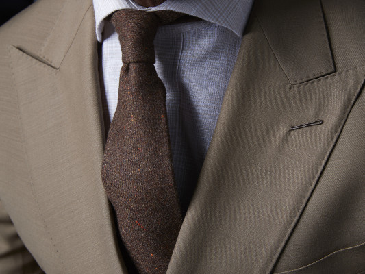 Link to Suits section. Close up view of a grey suit jacket, showing hand sewn details, a white shirt and dark tie.