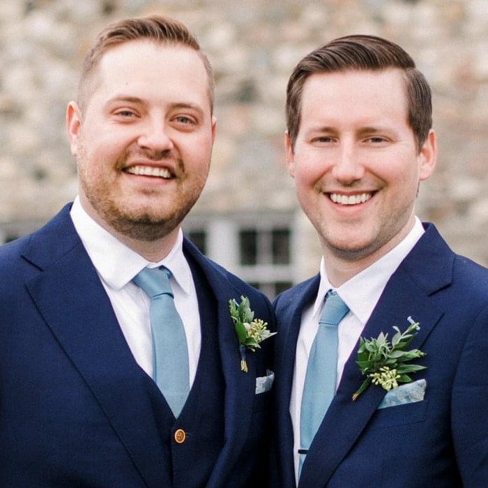 Two men wearing matching blue suits, white shirts, and light blue ties are smiling towards the camera.