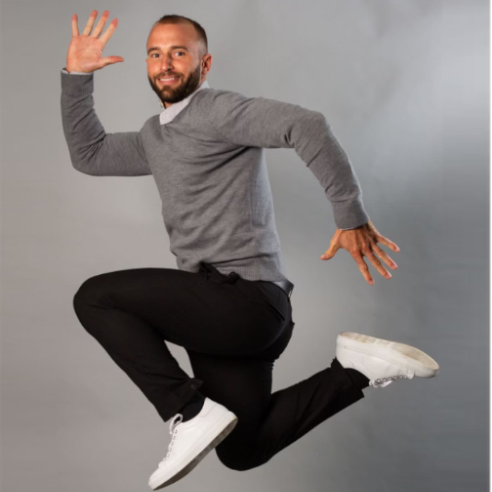 A man is jumping up against a grey background, showcasing his bright white shoes. He is wearing a grey sweater and black trousers.