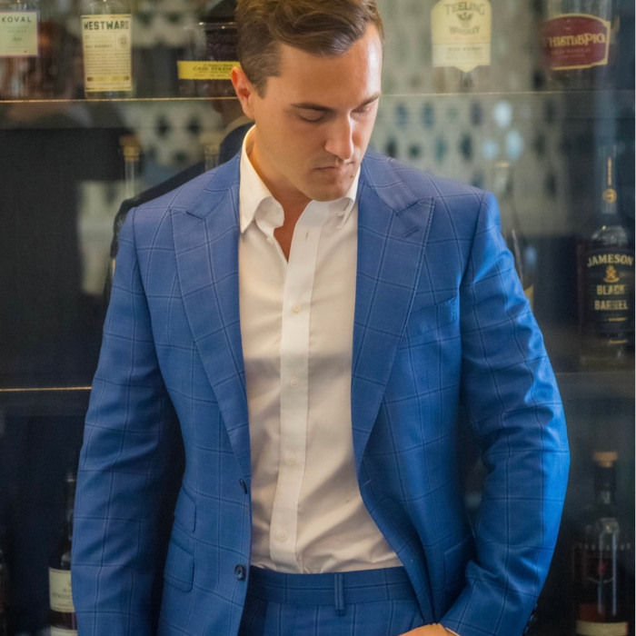 A man wearing a blue jacket and blue trousers and white shirt. He is standing in front of a bar