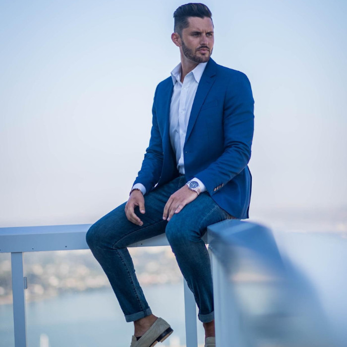 A man is sitting on the ledge of a balcony. He is wearing a blue suit, white shirt, and gray shoes.