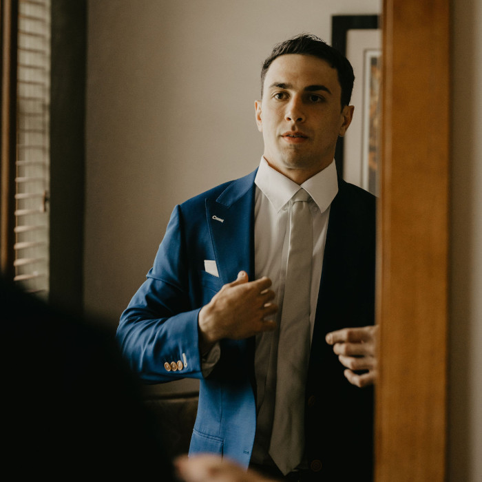 A man looking at his reflection in a mirror. He is adjusting his blue suit.