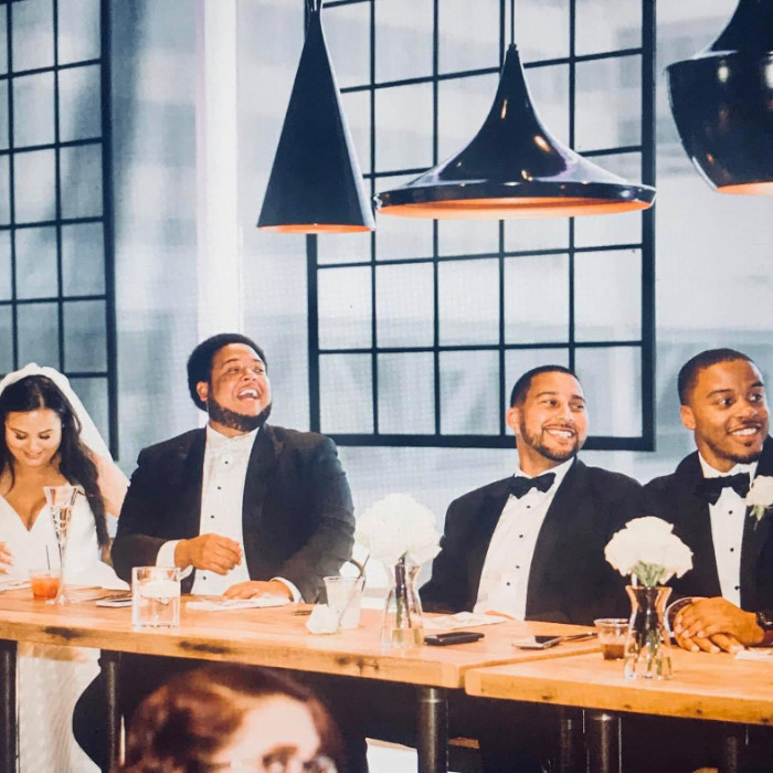 Three men wearing matching black tuxedos are sitting at a table with a woman in a white wedding dress. They are all smiling and looking out of the frame.