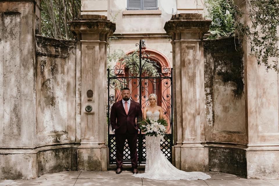 A wide shot of a bride and groom wedding portrait showing the groom in a well fitting burgundy suit. He is avoiding suit mistakes.